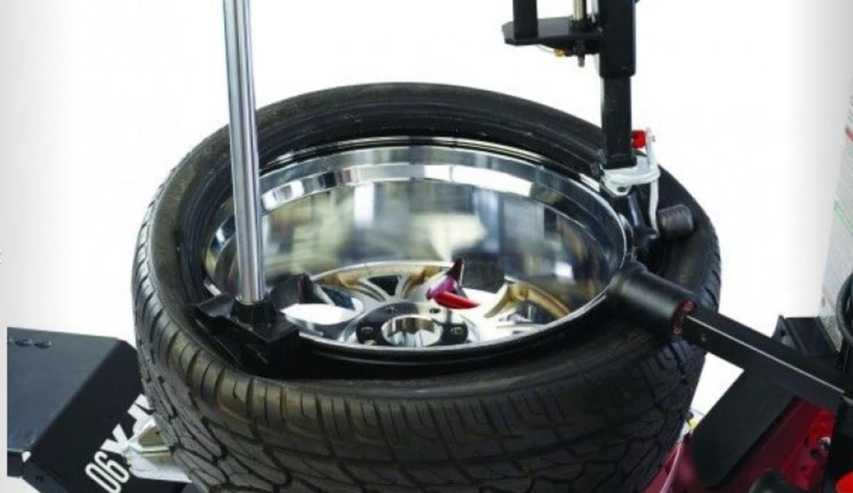 A tire is on the tabletop, while a roboarm and robo roller are pushing the tire down and around the wheel.