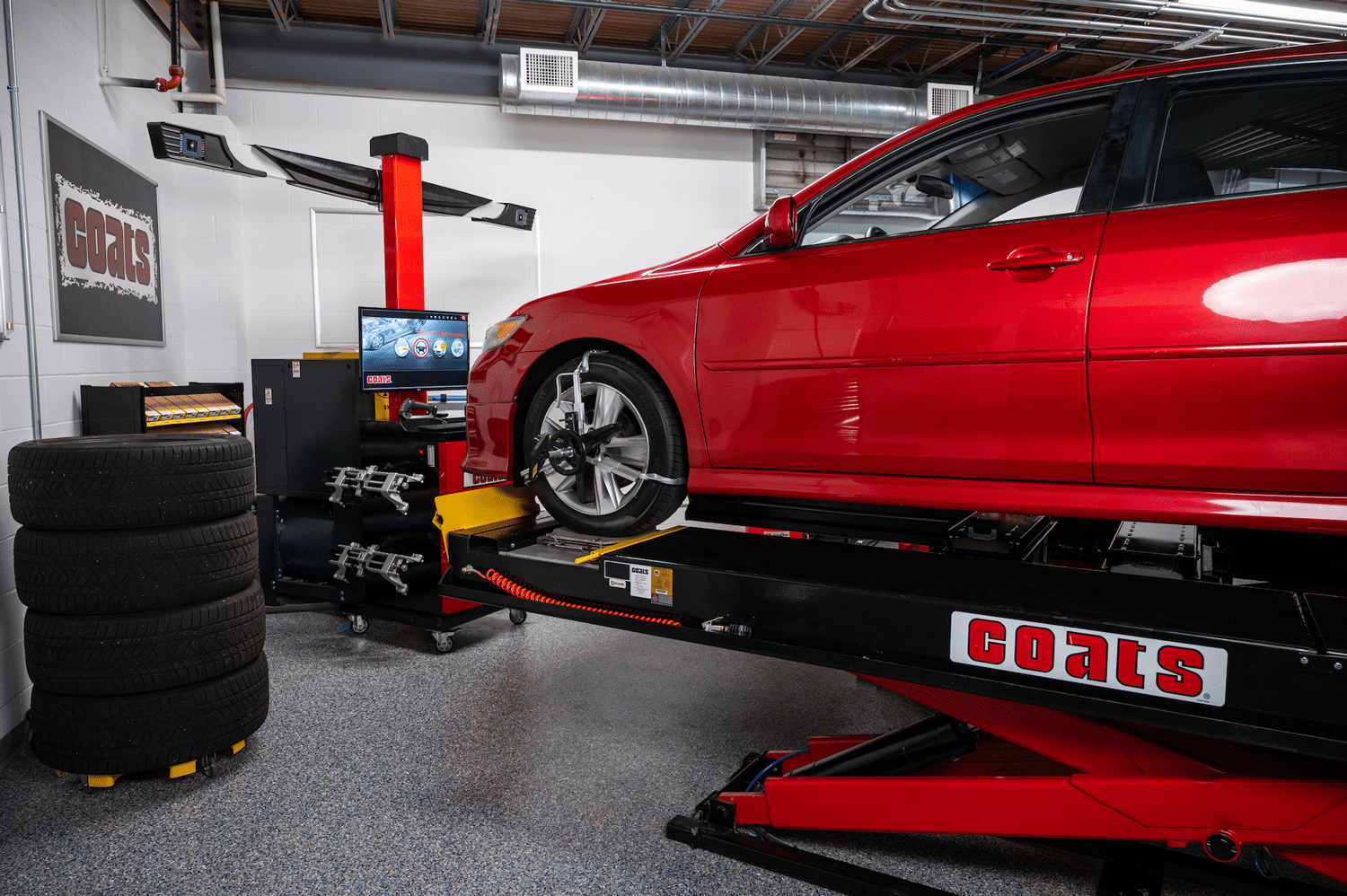 A red sedan is being serviced on a Coats alignment scissor lift, facing a Coats 6500 wheel aligner