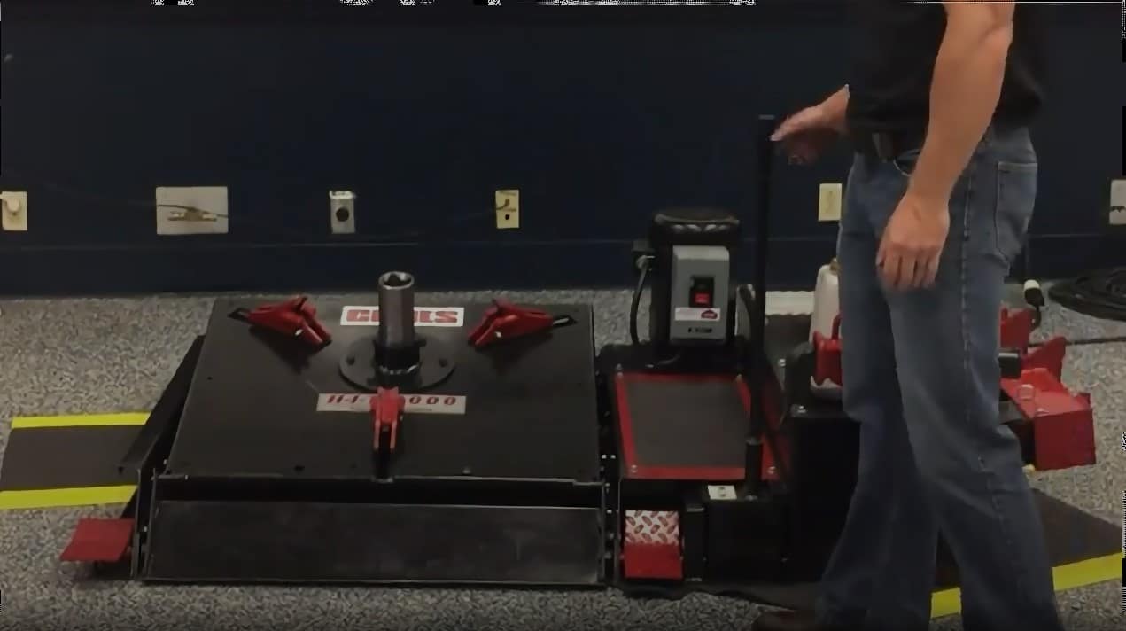 A technician uses a lever on the right of the HIT 6000 heavy duty tire changer to tilt up the tire changer platform, place the combo tool in the center, and then rotates the combo tool using foot pedals at the base of the machine.