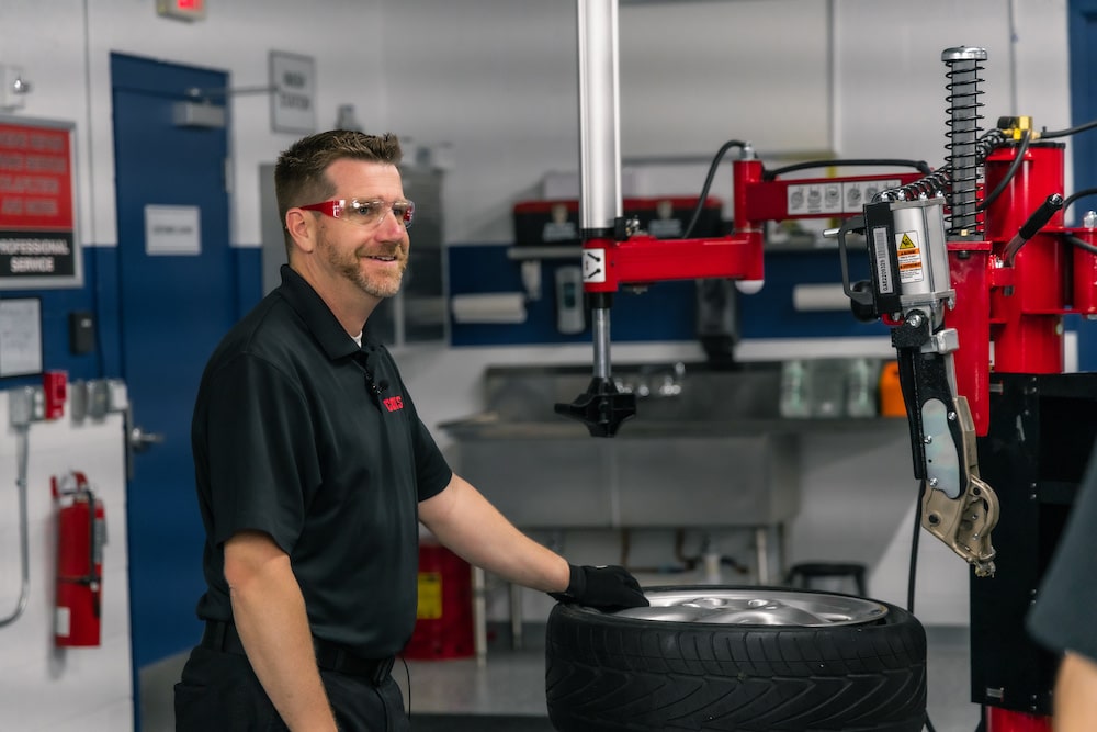 A Coats employee in a black shirt, work gloves, and safety glasses smiles as he changes a tire on a coats tire changer