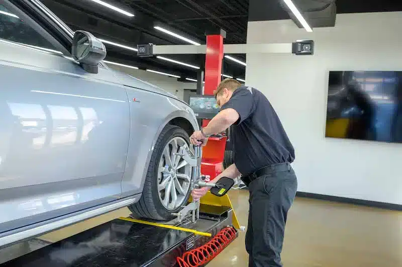 a man attaches a clamp to the wheel of a silver sedan in a shop with a CWA 6000 aligner